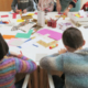 Des enfants sont assis autour d'une table recouverte de papier coloré, de bâtons de colle et de matériel de bricolage. Ils participent à une activité de bricolage au musée de la Toile de Jouy. Des adultes les aident et les guident. - Musée de la Toile de Jouy