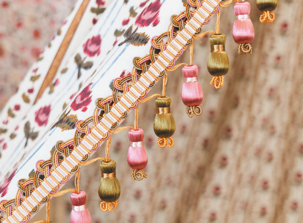 Gros plan sur une garniture de tissu ornée de glands roses et vert olive suspendus à un bord décoratif tissé et doré. Inspiré par le musée de la Toile de Jouy, l'arrière-plan est constitué d'un tissu à motifs floraux dans les tons beige et rose. - Musée de la Toile de Jouy