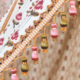 Gros plan sur une garniture de tissu ornée de glands roses et vert olive suspendus à un bord décoratif tissé et doré. Inspiré par le musée de la Toile de Jouy, l'arrière-plan est constitué d'un tissu à motifs floraux dans les tons beige et rose. - Musée de la Toile de Jouy