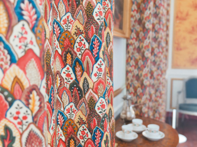 Gros plan sur un tissu coloré à motifs inspirés du Musée de la Toile de Jouy, présentant des feuilles et des motifs floraux. En arrière-plan flou, une table en bois contient des tasses à thé blanches, des soucoupes et une théière en métal dans une pièce confortable et élégante. - Musée de la Toile de Jouy