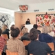 Une femme, debout sur un podium, s'adresse à un public assis dans une salle du musée de la Toile de Jouy, décorée de motifs floraux et de papiers peints en noir et blanc. Les personnes présentes écoutent attentivement. - Musée de la Toile de Jouy