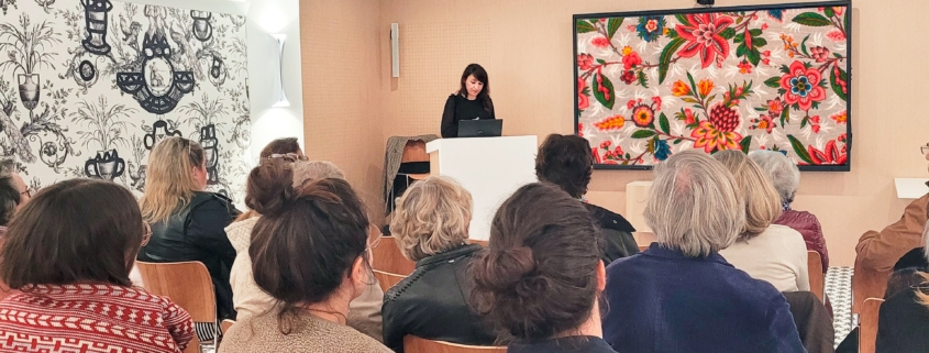 Une femme, debout sur un podium, s'adresse à un public assis dans une salle du musée de la Toile de Jouy, décorée de motifs floraux et de papiers peints en noir et blanc. Les personnes présentes écoutent attentivement. - Musée de la Toile de Jouy