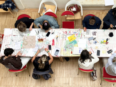 Au musée de la Toile de Jouy, un groupe de personnes est assis autour d'une table couverte de fournitures d'écriture et d'art colorées, travaillant sur des projets créatifs. La photo, prise du dessus, montre des outils, du papier et des cahiers éparpillés sur la table. - Musée de la Toile de Jouy