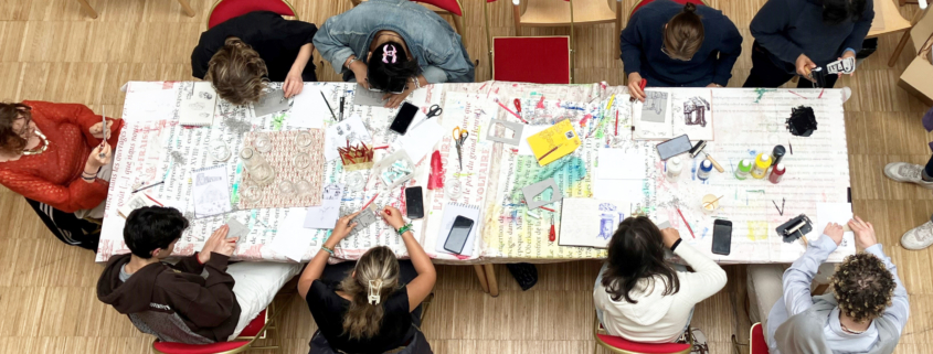 Au musée de la Toile de Jouy, un groupe de personnes est assis autour d'une table couverte de fournitures d'écriture et d'art colorées, travaillant sur des projets créatifs. La photo, prise du dessus, montre des outils, du papier et des cahiers éparpillés sur la table. - Musée de la Toile de Jouy