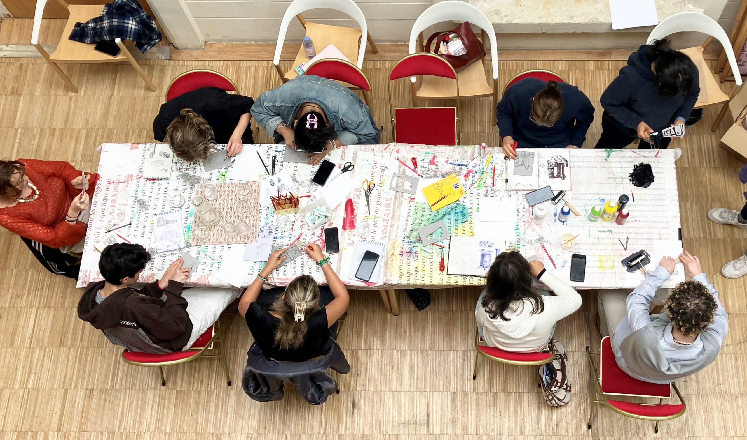 Au musée de la Toile de Jouy, un groupe de personnes est assis autour d'une table couverte de fournitures d'écriture et d'art colorées, travaillant sur des projets créatifs. La photo, prise du dessus, montre des outils, du papier et des cahiers éparpillés sur la table. - Musée de la Toile de Jouy