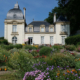 Un château historique avec deux tours arrondies, abritant le musée de la Toile de Jouy, se trouve derrière des jardins de fleurs colorés, entouré de verdure sous un ciel bleu clair. - Musée de la Toile de Jouy