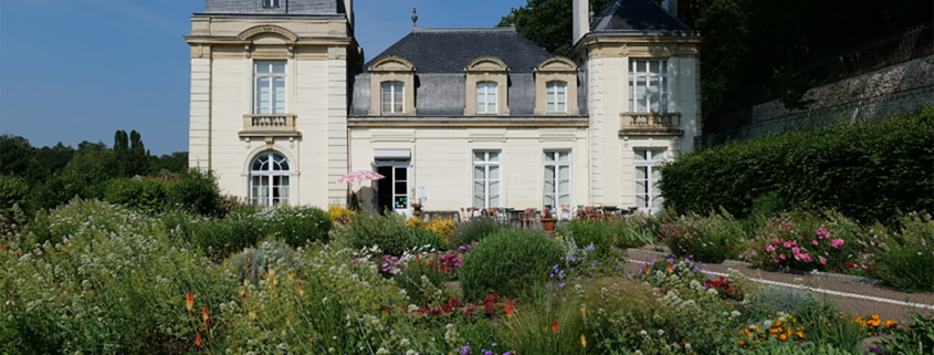 Un château historique avec deux tours arrondies, abritant le musée de la Toile de Jouy, se trouve derrière des jardins de fleurs colorés, entouré de verdure sous un ciel bleu clair. - Musée de la Toile de Jouy