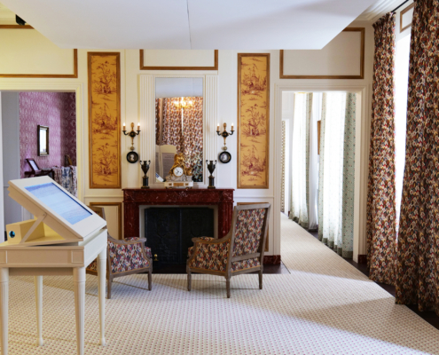 Une salle de style ancien du musée de la Toile de Jouy avec des fauteuils à motifs, une cheminée avec horloge et candélabres, des murs dorés, des rideaux fleuris et un présentoir d'information au premier plan. - Musée de la Toile de Jouy