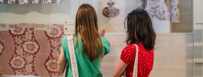 Deux femmes se tiennent devant une exposition du Musée de la Toile de Jouy présentant des tissus à motifs, un collier et un chemisier historique sur un mannequin, examinant attentivement l'exposition. - Musée de la Toile de Jouy