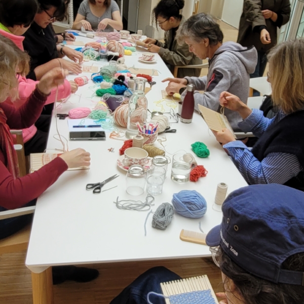 Un groupe de personnes est assis autour d'une longue table blanche au Musée de la Toile de Jouy, en train de tisser et de travailler avec des fils colorés. La table est recouverte de matériel de bricolage comme des ciseaux, des bobines et des bocaux dans ce cadre lumineux et créatif. - Musée de la Toile de Jouy