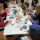 Un groupe de personnes est assis autour d'une longue table blanche au Musée de la Toile de Jouy, en train de tisser et de travailler avec des fils colorés. La table est recouverte de matériel de bricolage comme des ciseaux, des bobines et des bocaux dans ce cadre lumineux et créatif. - Musée de la Toile de Jouy