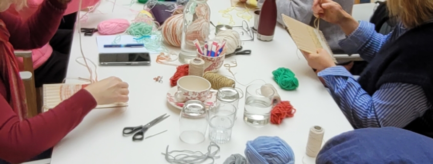 Un groupe de personnes est assis autour d'une longue table blanche au Musée de la Toile de Jouy, en train de tisser et de travailler avec des fils colorés. La table est recouverte de matériel de bricolage comme des ciseaux, des bobines et des bocaux dans ce cadre lumineux et créatif. - Musée de la Toile de Jouy