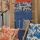 Exposition de textiles à motifs, principalement bleus et orange, drapés sur des étagères en bois au Musée de la Toile de Jouy. Les dessins représentent des scènes et des motifs complexes (personnages, arbres, motifs géométriques), tandis que la lumière filtre à travers une fenêtre voisine. - Musée de la Toile de Jouy
