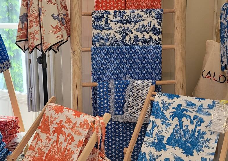 Exposition de textiles à motifs, principalement bleus et orange, drapés sur des étagères en bois au Musée de la Toile de Jouy. Les dessins représentent des scènes et des motifs complexes (personnages, arbres, motifs géométriques), tandis que la lumière filtre à travers une fenêtre voisine. - Musée de la Toile de Jouy