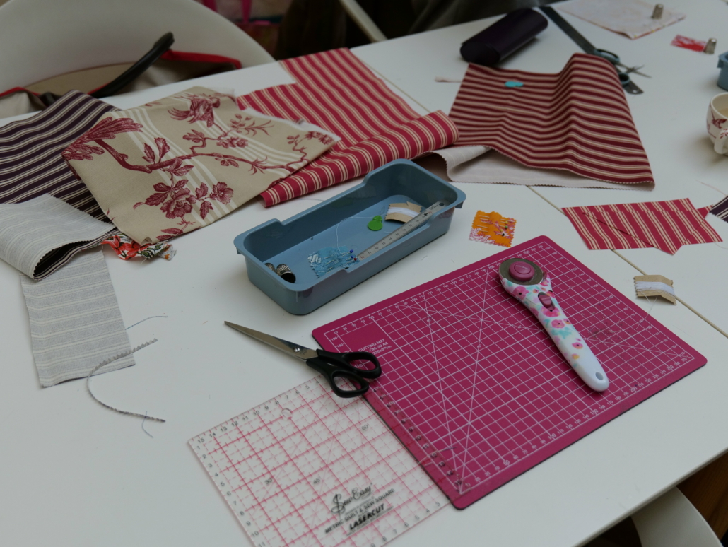 Une table blanche présente des pièces de tissu à rayures et à fleurs, des ciseaux, un cutter rotatif sur un tapis de coupe rose, une règle transparente et un plateau bleu avec des épingles à coudre - évoquant l'art textile intemporel du Musée de la Toile de Jouy. - Musée de la Toile de Jouy