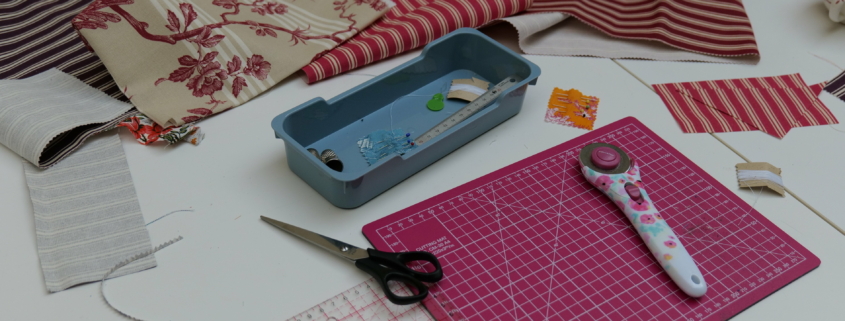 Une table blanche présente des pièces de tissu à rayures et à fleurs, des ciseaux, un cutter rotatif sur un tapis de coupe rose, une règle transparente et un plateau bleu avec des épingles à coudre - évoquant l'art textile intemporel du Musée de la Toile de Jouy. - Musée de la Toile de Jouy