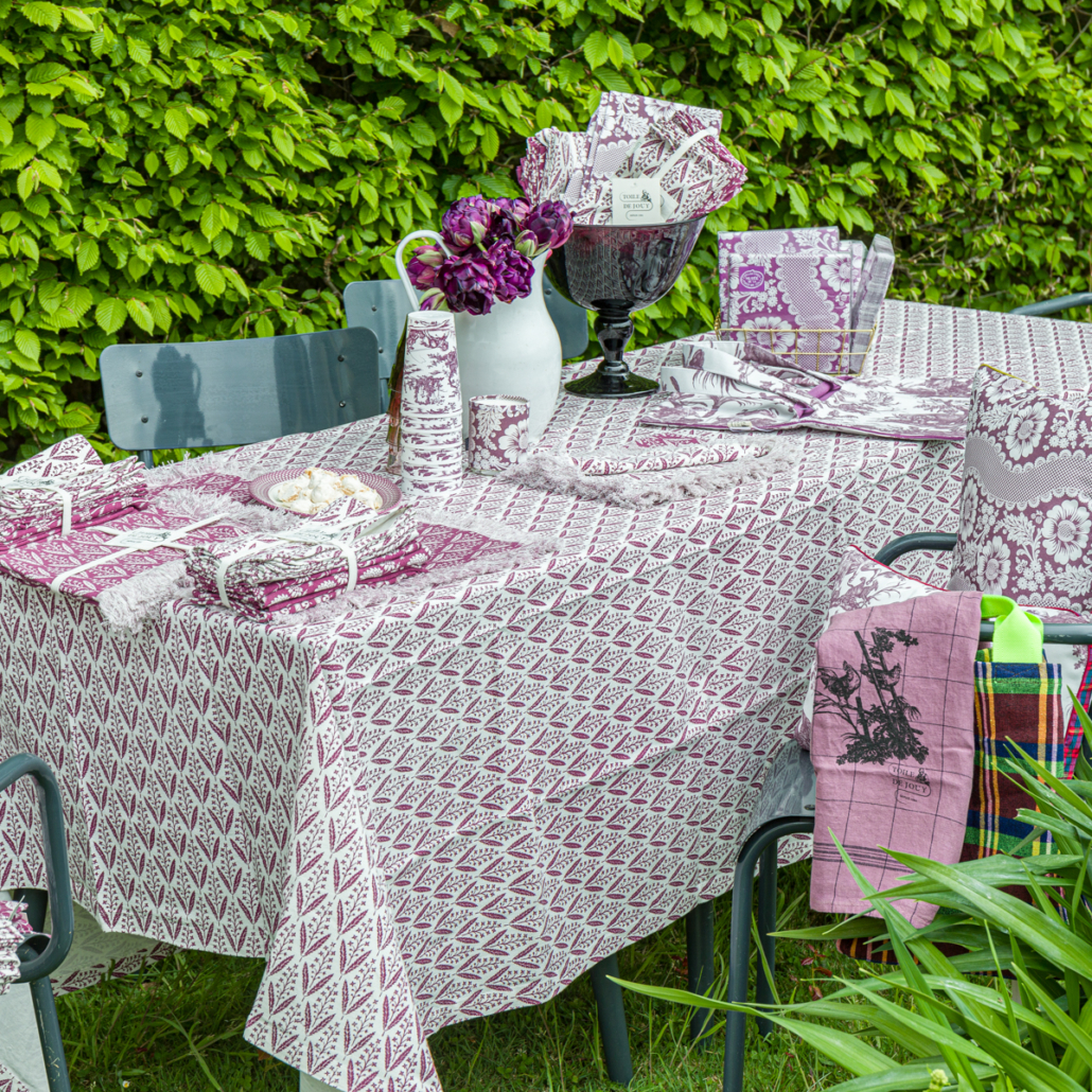 Une table de jardin dressée pour un repas avec une nappe à motifs violets et blancs, des serviettes et de la vaisselle du Marché de l'Eglantine. Un pichet blanc contenant des fleurs violettes et un bol avec des serviettes sont placés au centre ; des chaises vertes et une haie feuillue forment l'arrière-plan. - Musée de la Toile de Jouy