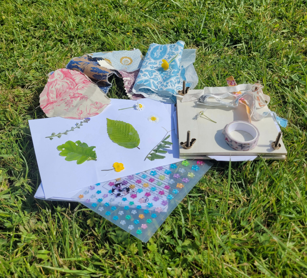 Une collection de matériel de bricolage, des feuilles pressées, des fleurs, des papiers à motifs et un petit journal avec du ruban et du washi tape du Marché de l'Eglantine, posés sur de l'herbe verte sous un soleil radieux. - Musée de la Toile de Jouy