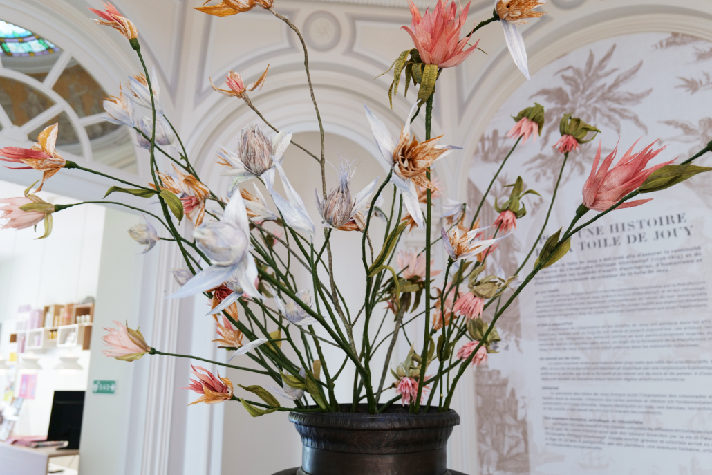 Un vase sombre rempli de fleurs artificielles roses et blanches est posé à l'intérieur contre un mur clair et orné de moulures décoratives, rappelant les présentoirs du marché de l'Églantine, avec un grand panneau d'information en arrière-plan. - Musée de la Toile de Jouy