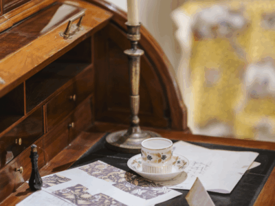 Un bureau à roulettes en bois, rappelant le Musée de la Toile de Jouy, contient des papiers épars avec des dessins, un chandelier avec une bougie éteinte, une tasse à thé et une soucoupe, une enveloppe et une petite pièce d'échecs pour une scène d'espace de travail vintage et confortable. - Musée de la Toile de Jouy