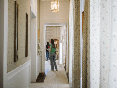 Un couloir au papier peint et à la moquette à motifs, rappelant le musée de la Toile de Jouy, mène à une pièce où se tiennent deux personnes, l'une en chemise verte et sac à dos, le regard partiellement détourné, l'autre près d'un meuble. - Musée de la Toile de Jouy