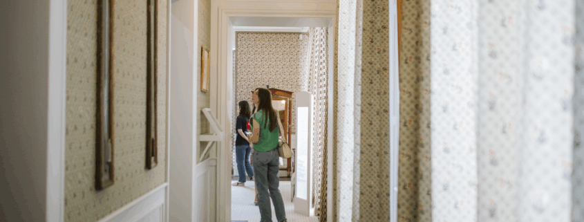Deux personnes se tiennent au fond d'un couloir lumineux, recouvert de moquette, dont le papier peint et les rideaux à motifs rappellent ceux du musée de la Toile de Jouy. L'une des personnes porte une chemise verte et un jean, ainsi qu'un sac beige. Des luminaires sont suspendus au plafond. - Musée de la Toile de Jouy