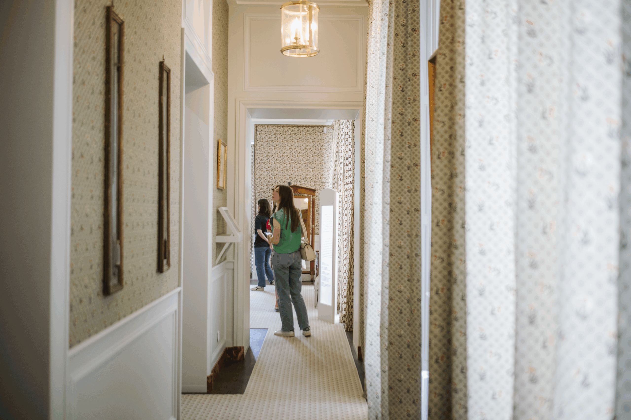 Deux personnes se tiennent au fond d'un couloir lumineux, recouvert de moquette, dont le papier peint et les rideaux à motifs rappellent ceux du musée de la Toile de Jouy. L'une des personnes porte une chemise verte et un jean, ainsi qu'un sac beige. Des luminaires sont suspendus au plafond. - Musée de la Toile de Jouy
