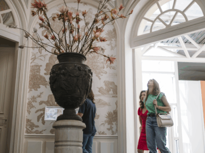 Trois personnes passent devant un grand vase orné de branches dans une grande salle à coupole du musée de la Toile de Jouy, avec des murs décoratifs, des fenêtres cintrées et la lumière naturelle qui pénètre par une porte ouverte. - Musée de la Toile de Jouy