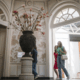 Trois personnes passent devant un grand vase orné de branches dans une grande salle à coupole du musée de la Toile de Jouy, avec des murs décoratifs, des fenêtres cintrées et la lumière naturelle qui pénètre par une porte ouverte. - Musée de la Toile de Jouy