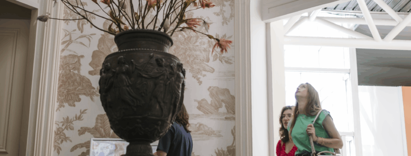 Trois personnes passent devant un grand vase orné de branches dans une grande salle à coupole du musée de la Toile de Jouy, avec des murs décoratifs, des fenêtres cintrées et la lumière naturelle qui pénètre par une porte ouverte. - Musée de la Toile de Jouy