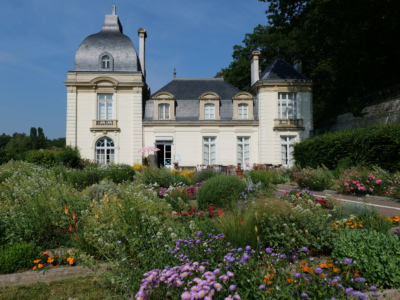 Un château de couleur claire avec deux tours à dôme, abritant le musée de la Toile de Jouy, se dresse derrière un jardin vibrant rempli de fleurs et de verdure variées sous un ciel bleu clair. - Musée de la Toile de Jouy