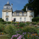 Un château de couleur claire avec deux tours à dôme, abritant le musée de la Toile de Jouy, se dresse derrière un jardin vibrant rempli de fleurs et de verdure variées sous un ciel bleu clair. - Musée de la Toile de Jouy