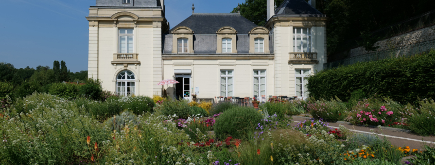 Un château de couleur claire avec deux tours à dôme, abritant le musée de la Toile de Jouy, se dresse derrière un jardin vibrant rempli de fleurs et de verdure variées sous un ciel bleu clair. - Musée de la Toile de Jouy