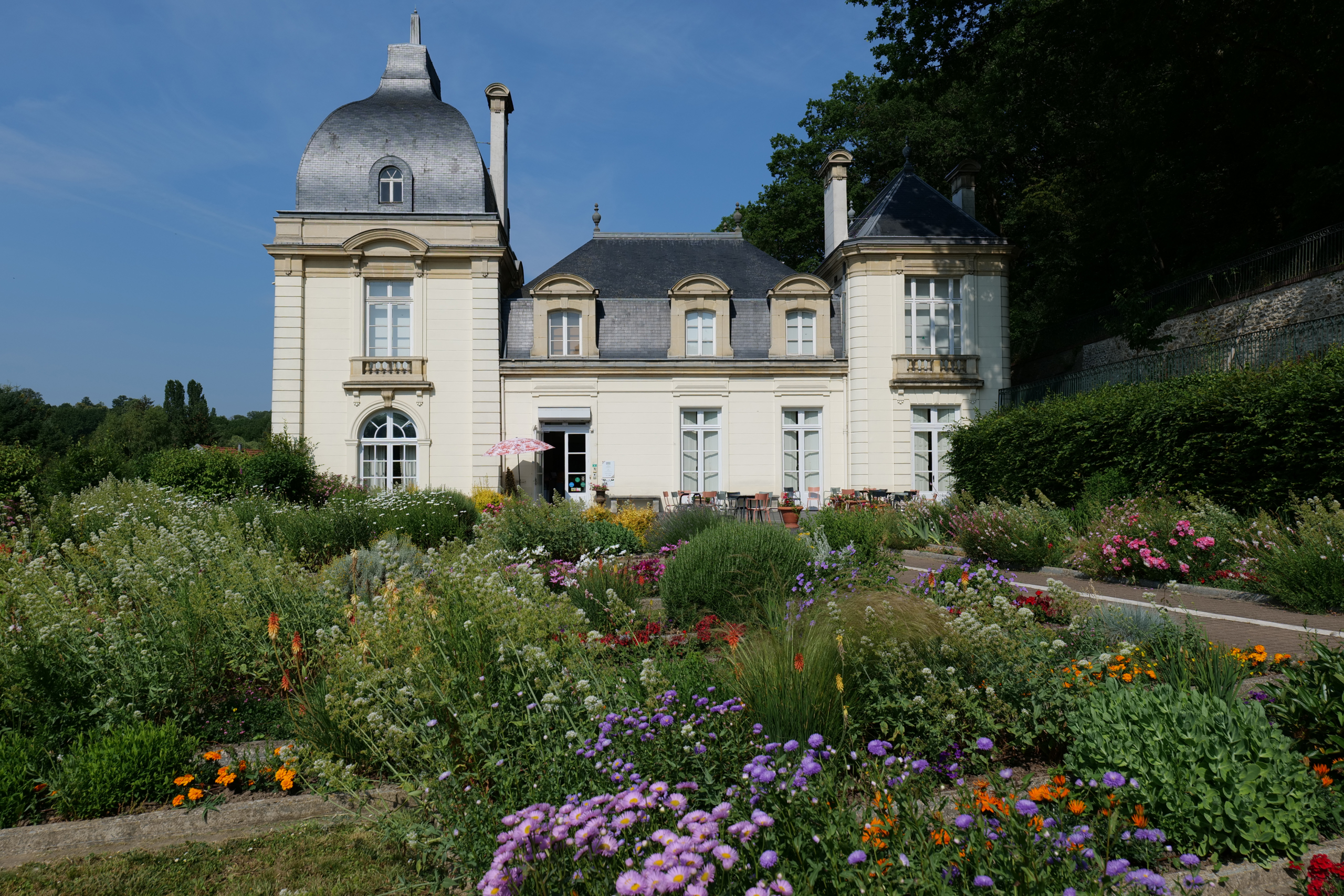 Un château de couleur claire avec deux tours à dôme, abritant le musée de la Toile de Jouy, se dresse derrière un jardin vibrant rempli de fleurs et de verdure variées sous un ciel bleu clair. - Musée de la Toile de Jouy