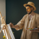 Un homme portant un chapeau de paille et une chemise beige ample peint à un chevalet dans une salle faiblement éclairée du musée de la Toile de Jouy. Il tient une palette contenant des peintures colorées et a de la peinture jaune sur le visage. - Musée de la Toile de Jouy