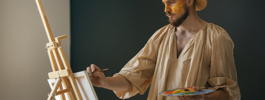 Un homme portant un chapeau de paille et une chemise beige ample peint à un chevalet dans une salle faiblement éclairée du musée de la Toile de Jouy. Il tient une palette contenant des peintures colorées et a de la peinture jaune sur le visage. - Musée de la Toile de Jouy
