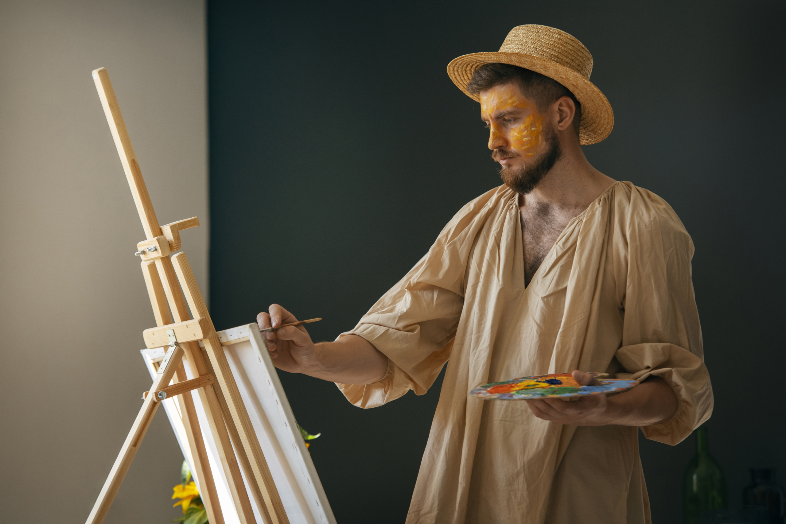 Un homme portant un chapeau de paille et une chemise beige ample peint à un chevalet dans une salle faiblement éclairée du musée de la Toile de Jouy. Il tient une palette contenant des peintures colorées et a de la peinture jaune sur le visage. - Musée de la Toile de Jouy