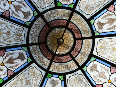 Un dôme en vitrail du musée de la Toile de Jouy, aux motifs floraux et géométriques complexes, présente des jaunes pâles, des roses, des verts et des bleus rayonnant à partir d'un motif circulaire central. Des lignes noires divisent les sections. - Musée de la Toile de Jouy