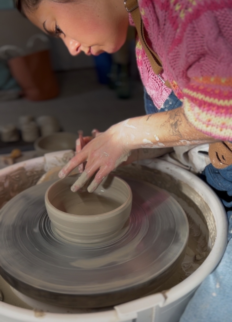 Au Marché de l'Eglantine, une personne vêtue d'un pull rose façonne de l'argile sur un tour de poterie, se concentrant sur la formation d'un bol rond tandis que le tour tourne et que l'argile éclabousse le poste de travail occupé. - Musée de la Toile de Jouy