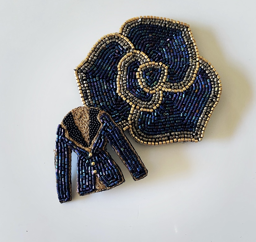 Deux broches en perles sur fond blanc : l'une est une fleur stylisée bleu et or, l'autre est une veste détaillée bleu et or - des pièces uniques inspirées par le charme du Marché de l'Eglantine. - Musée de la Toile de Jouy