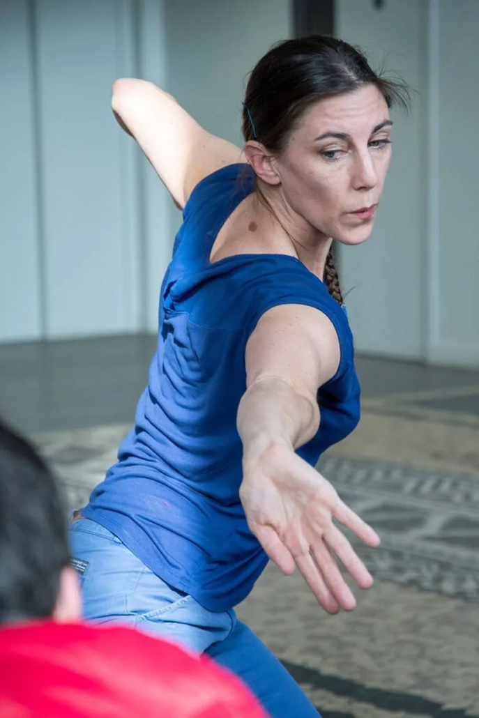 Une femme vêtue d'une chemise bleue et d'un jean tend gracieusement le bras vers l'avant tout en dansant à l'intérieur du Marché de l'Eglantine, son expression étant concentrée et intentionnelle. Une autre personne en chemise rouge est partiellement visible au premier plan. - Musée de la Toile de Jouy