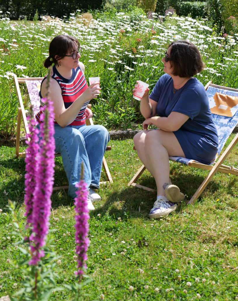 Deux femmes sont assises sur des chaises longues dans un jardin herbeux, discutant et buvant des boissons. Entourées de fleurs sauvages, dont des marguerites et de grandes fleurs roses, elles profitent d'une journée ensoleillée en plein air au Marché de l'Églantine. - Musée de la Toile de Jouy