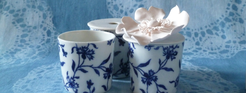 Trois tasses en porcelaine blanche à motifs floraux bleus sont présentées sur un tissu bleu à motifs de dentelle, évoquant l'élégance raffinée du musée de la Toile de Jouy. L'une des tasses est surmontée d'une délicate fleur blanche. - Musée de la Toile de Jouy