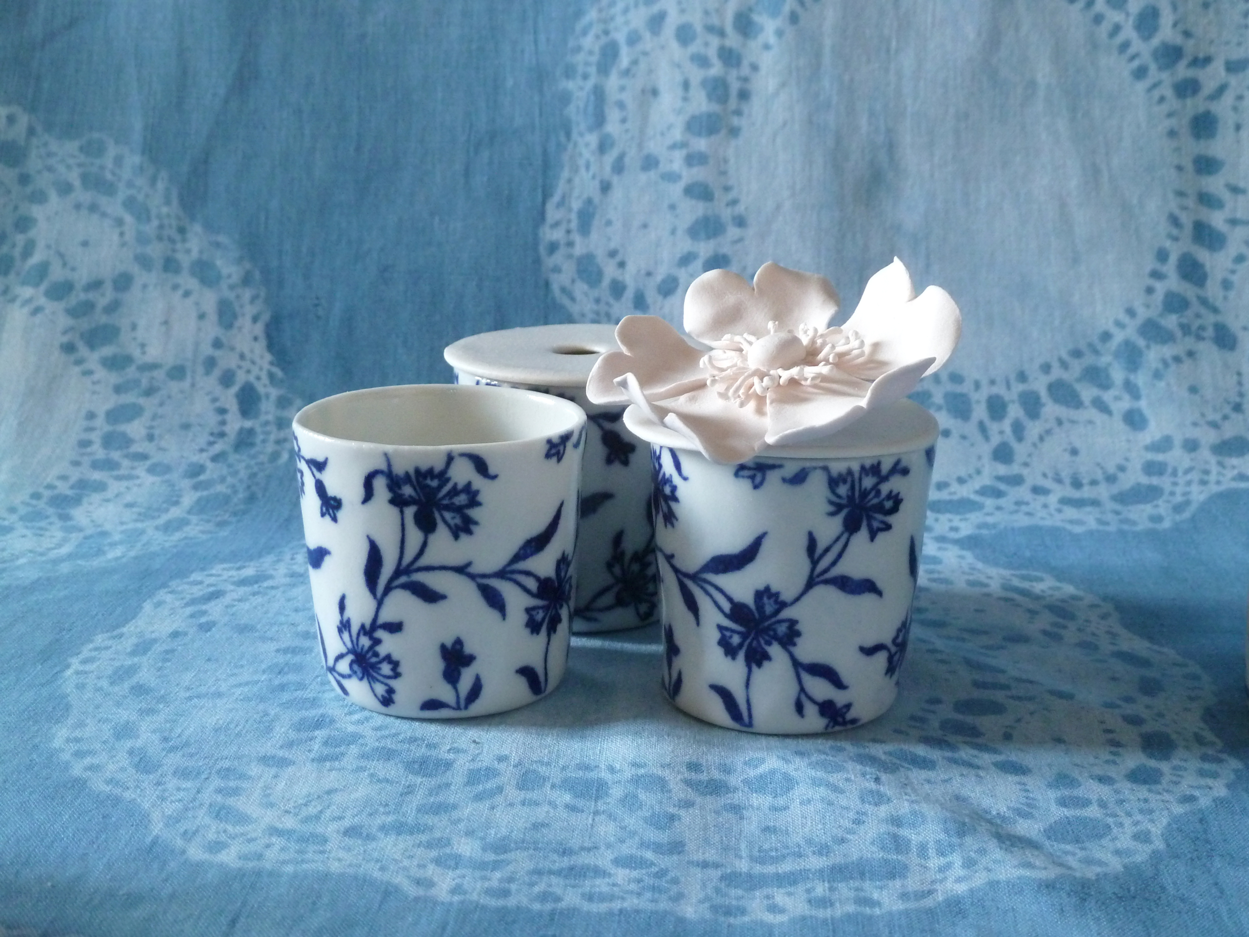 P1340248_ Trois tasses en porcelaine blanche à motifs floraux bleus sont présentées sur un tissu bleu à motifs de dentelle, évoquant l'élégance raffinée du musée de la Toile de Jouy. L'une des tasses est surmontée d'une délicate fleur blanche. - Musée de la Toile de Jouy