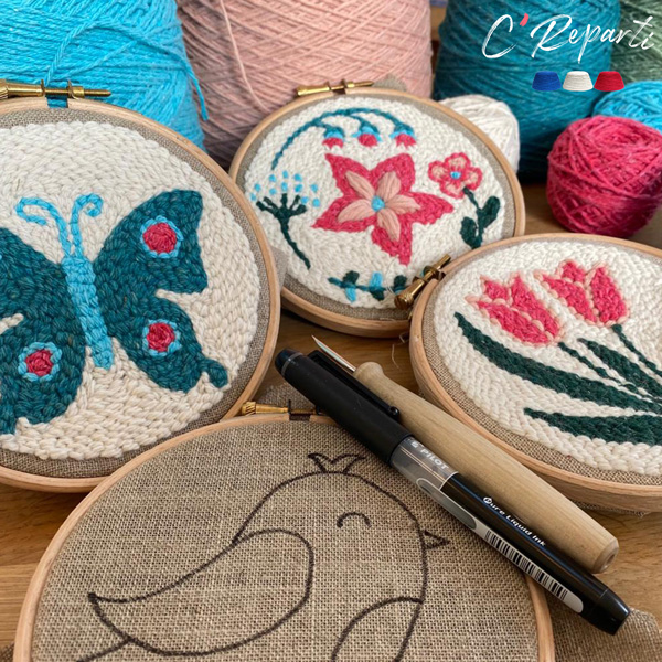 Trois cercles de broderie présentent des motifs colorés : un papillon, des fleurs et une tulipe. L'un des cercles présente un croquis d'oiseau inachevé. Des stylos et des pelotes de laine sont posés à proximité - une inspiration parfaite pour le marché de l'Eglantine. - Musée de la Toile de Jouy