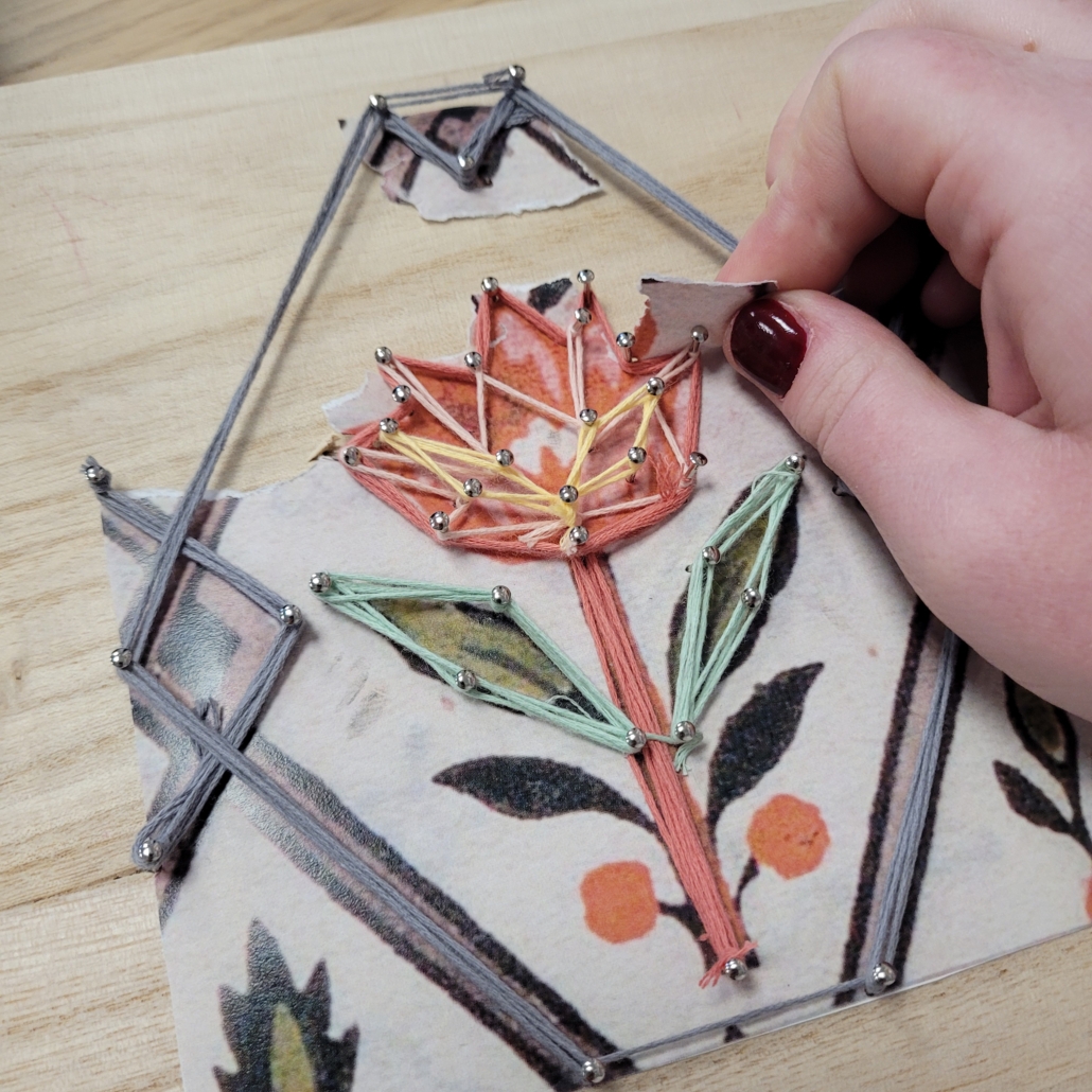Une main utilise un petit outil pour guider un fil coloré autour de clous sur une planche en bois, créant ainsi une fleur de string art sur un dessin imprimé comme référence - faisant écho aux traditions créatives trouvées au Musée de la Toile de Jouy. - Musée de la Toile de Jouy