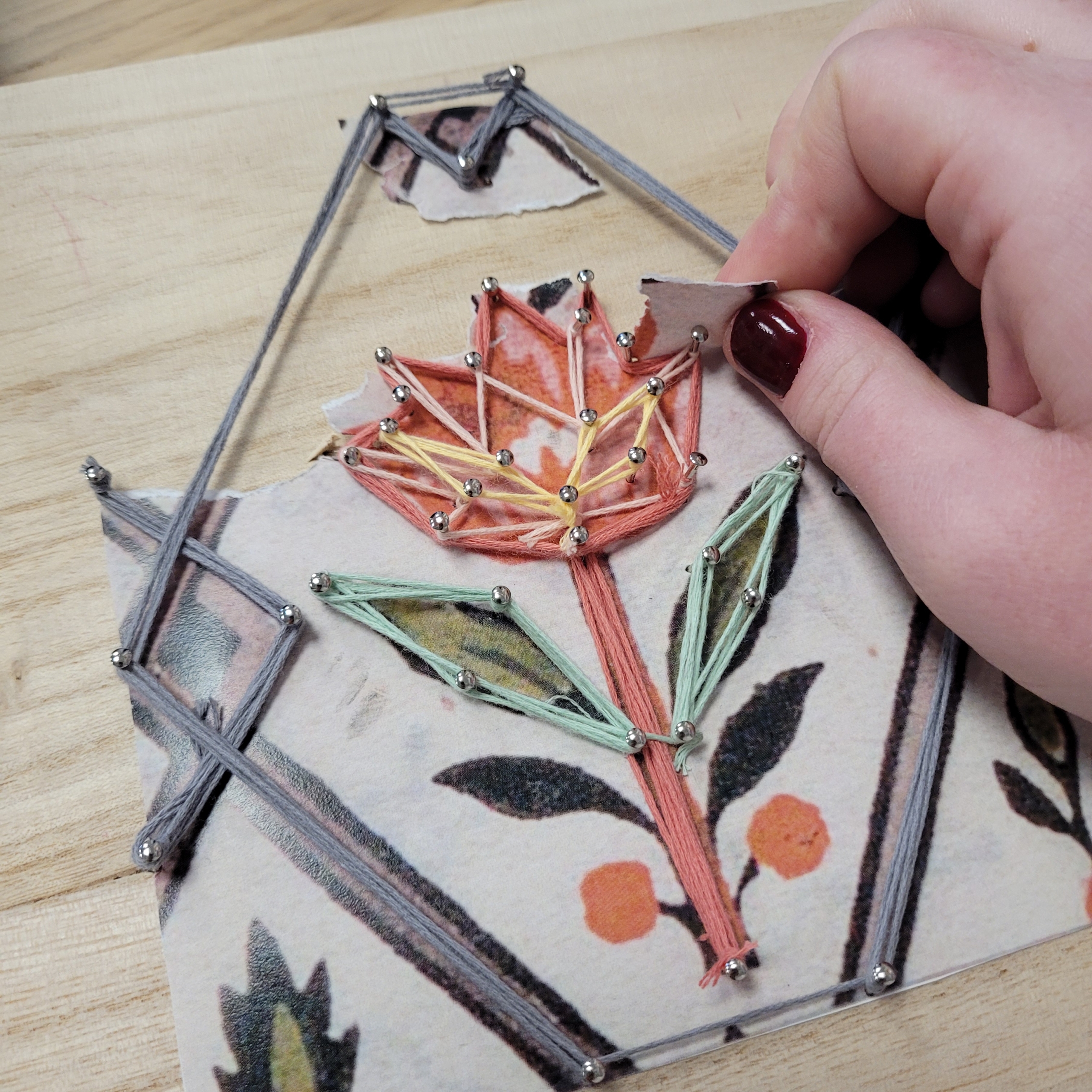 Une main utilise un petit outil pour guider un fil coloré autour de clous sur une planche en bois, créant ainsi une fleur de string art sur un dessin imprimé comme référence - faisant écho aux traditions créatives trouvées au Musée de la Toile de Jouy. - Musée de la Toile de Jouy