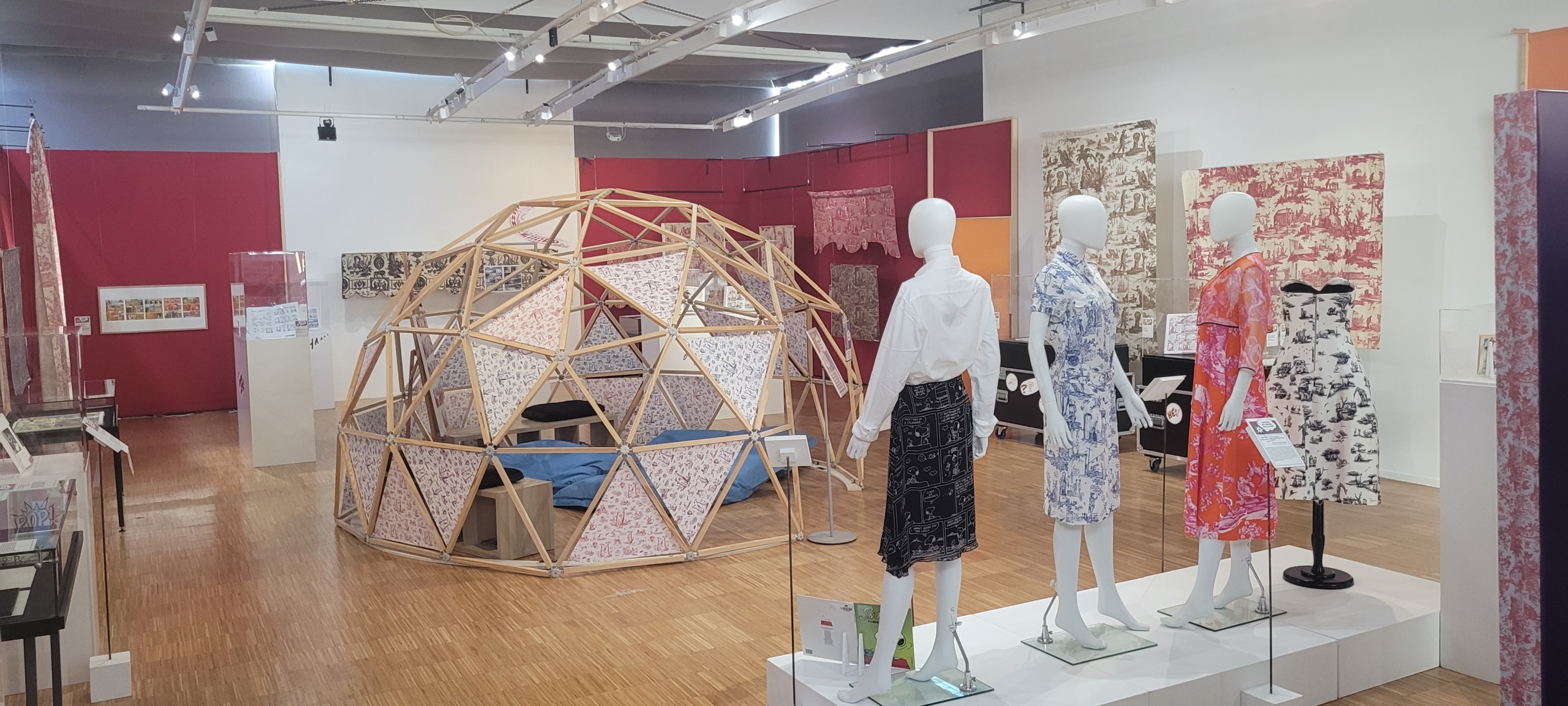 Une salle d'exposition d'art moderne présente des mannequins vêtus de robes à motifs colorés, une structure de dôme géodésique en bois et des œuvres d'art textile exposées sur les murs et les stands. - Musée de la Toile de Jouy