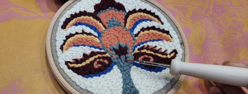 Une main tient une aiguille à poinçonner, travaillant sur un cercle de broderie avec un motif floral ou abstrait coloré dans un fil marron, bleu, orange et crème. Dans cet atelier confortable, une pelote de laine est posée à proximité sur la surface à motifs dorés. - Musée de la Toile de Jouy