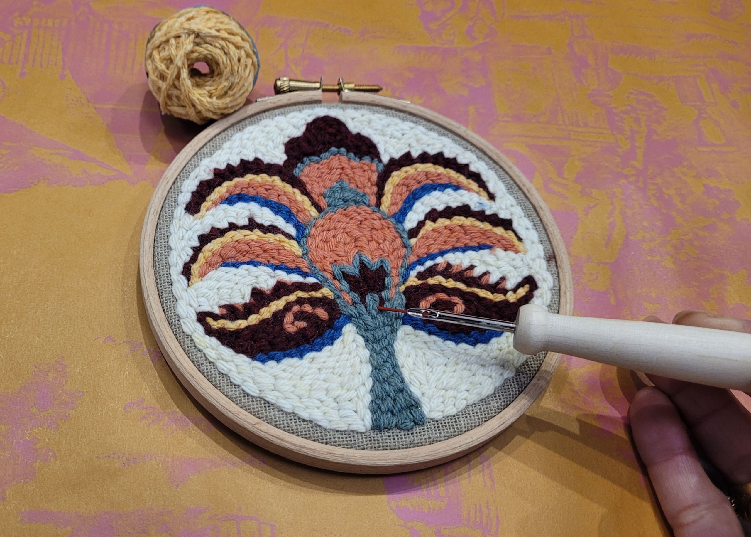 Une main tient une aiguille à poinçonner, travaillant sur un cercle de broderie avec un motif floral ou abstrait coloré dans un fil marron, bleu, orange et crème. Dans cet atelier confortable, une pelote de laine est posée à proximité sur la surface à motifs dorés. - Musée de la Toile de Jouy